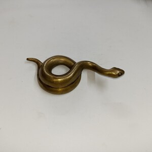May include: A coiled brass snake figurine with a detailed head and body. The snake is a golden-brown colour and appears to be made of metal. The figurine is on a white surface.