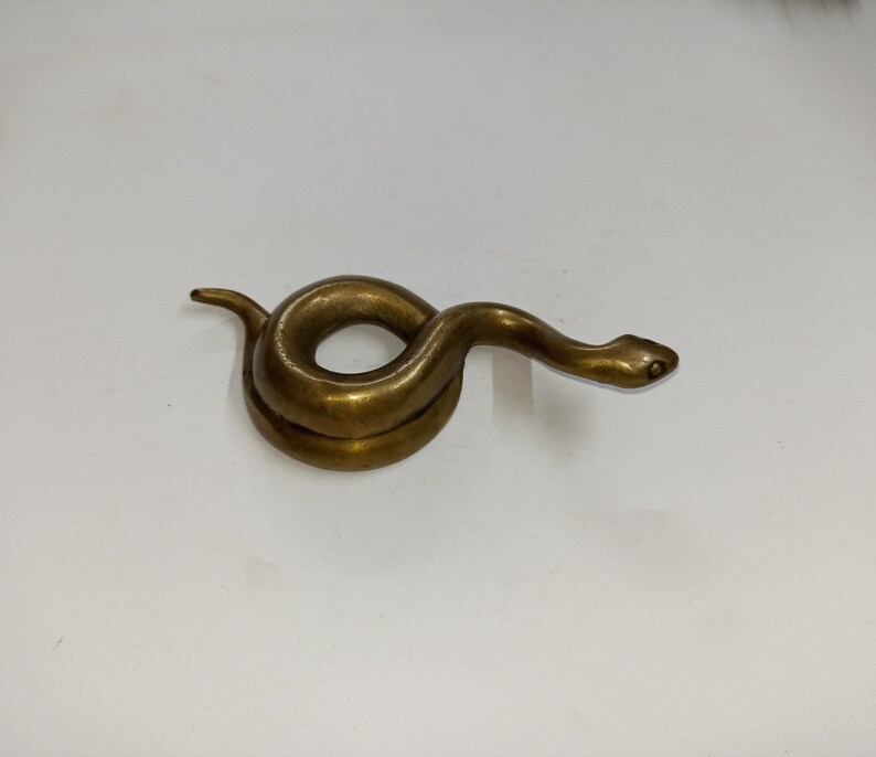 May include: A coiled brass snake figurine with a detailed head and body. The snake is a golden-brown colour and appears to be made of metal. The figurine is on a white surface.