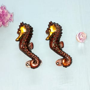 May include: Two bronze seahorse-shaped cabinet pulls with a gold-coloured head and a reddish-brown body. The seahorses are mounted on a light blue surface, with a pink flower and a decorative knob in the background.