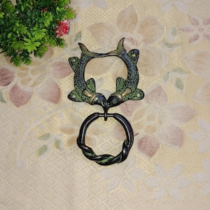May include: A decorative metal door knocker featuring two fish facing each other, forming a circular opening. The fish are detailed with green and gold accents. Below, a twisted ring handle completes the design.