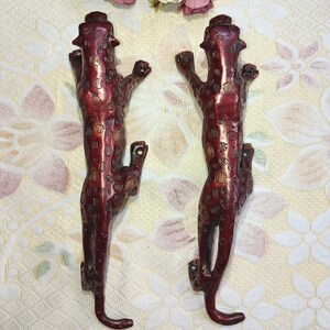 Könnte beinhalten: Zwei rotbraune Türgriffe in Leopardenform. Die Griffe sind mit Flecken versehen und vertikal montiert. Sie bestehen aus Metall und haben eine Vintage-Ästhetik. Der Hintergrund ist ein Stoff mit Blumenmuster.