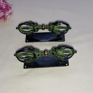 Op de afbeelding: Een paar zwarte en groene metalen deurgrepen met een decoratief ontwerp. De grepen zijn gemonteerd op zwarte rechthoekige platen.