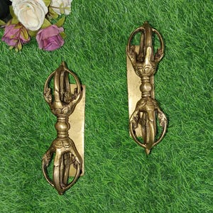 May include: Two gold-coloured metal Tibetan ritual objects with intricate designs, mounted on rectangular wooden plaques. The objects are shaped like a vajra, a symbolic weapon in Buddhism.