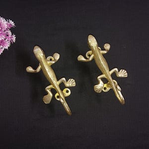 Peut inclure: Deux poignées de meubles dorées en forme de lézard sur une surface noire. Les poignées présentent des écailles texturées et une queue en forme de crochet. Une grappe de fleurs roses est dans le coin supérieur gauche. Ces poignées décoratives ajoutent une touche unique aux meubles.