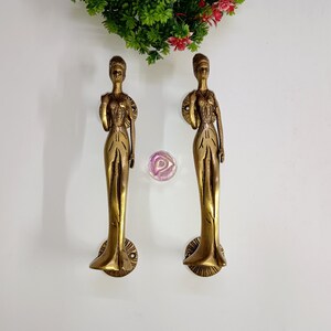 May include: A pair of gold-toned door handles shaped like standing figures. The handles are detailed with intricate designs and have a smooth, polished finish.