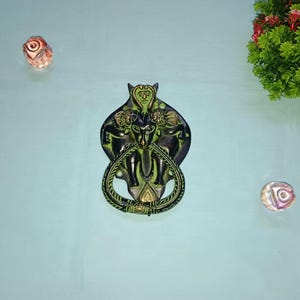 Könnte beinhalten: Ein grüner und schwarzer Türklopfer aus Metall mit einem detaillierten Design einer mythischen Kreatur mit Flügeln und Hörnern. Der Türklopfer hat einen Seilring zum Ziehen.