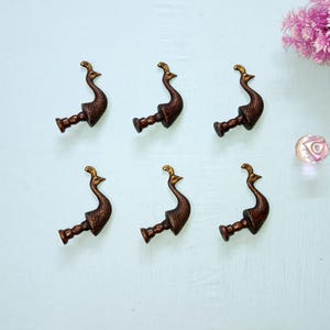 May include: Six bronze peacock-shaped drawer pulls with gold detailing. The pulls are displayed on a light blue surface, with a cluster of pink flowers and a clear glass knob in the background. Each pull is approximately 10 cm long.