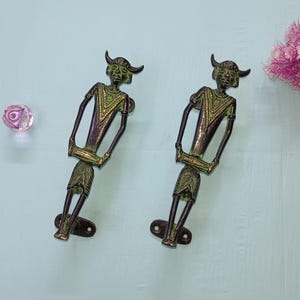 May include: Two bronze-colored door handles in the shape of stylized figures with horns, holding a drum-like object. The figures have green and gold accents. A pink and purple glass knob and a bouquet of pink flowers are also visible.