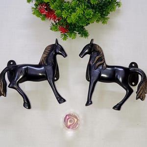 May include: Two black horse-shaped wall decorations with bronze manes and tails. The horse figures are mounted on a white wall, with a decorative floral arrangement above and a decorative crystal ball below.