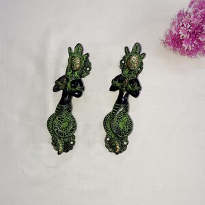 May include: Two ornate brass door handles with a green and black finish. Each handle features a female figure with a raised hand in a prayer position. The figures are adorned with intricate details and are set against a plain white background.