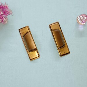 Könnte beinhalten: Zwei rechteckige Schubladengriffe aus Messing mit einem geschwungenen Mittelgriff. Die Griffe haben eine warme, gealterte Oberfläche, die eine Vintage-Ästhetik suggeriert. Der Hintergrund ist eine hellblaue Oberfläche mit einem rosa Blumenarrangement und einem dekorativen Glasobjekt.