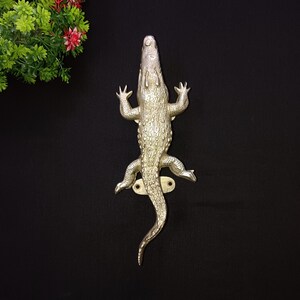 以下が含まれることがあります： 銀色のワニの形をしたドアハンドル。ハンドルは質感のある鱗で詳細に表現されており、尾と4本の脚があります。ハンドルは、黒い背景に対して真鍮色のプレートに取り付けられています。