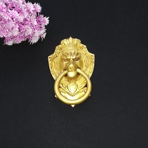 May include: A gold-colored lion head door knocker with a ring handle. The lion has a crown and a heart-shaped detail below the ring. The background is black, and a bouquet of pink flowers is in the upper left corner.
