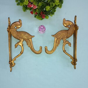 Pode incluir: Um par de ganchos de parede de metal dourado em forma de pavão. Os ganchos são ornamentados e detalhados, com as penas e as caudas dos pavões esculpidas de forma intrincada.