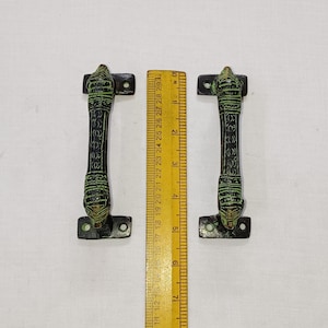 May include: Two antique bronze door handles with a dark green patina. Each handle is approximately 15 cm long, featuring an ornate design with decorative details. The handles are placed next to a wooden ruler.