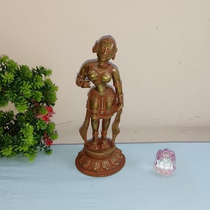 Puede incluir: Una estatua de color bronce de una mujer con ropa y joyas detalladas se encuentra sobre una base decorativa. La estatua está colocada junto a un pequeño objeto de vidrio facetado rosa y una planta verde con flores rojas y rosas.