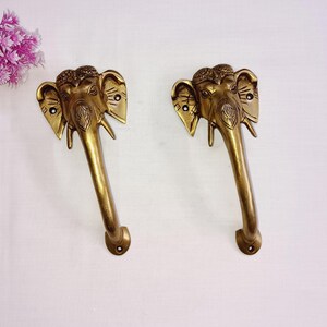 May include: Two gold-toned door handles shaped like elephant heads with intricate details. The handles have a long, curved design and are attached to a wall.