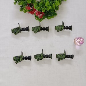 Può includere: Sei pomelli decorativi in metallo verde e nero con un design dettagliato a testa di drago. I pomelli sono disposti su una superficie bianca, con un piccolo oggetto di vetro sfaccettato rosa e vegetazione sullo sfondo.