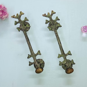 Może przedstawiać: Dwa brązowe uchwyty do drzwi z motywem gałęzi drzewa. Uchwyty mają długi, smukły trzon z teksturowanym wzorem i szczytem w kształcie drzewa. Uchwyty są zaakcentowane zielonymi i złotymi detalami. Różowy kwiat i różowa szklana kulka znajdują się w tle.