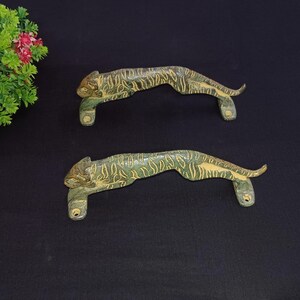 Peut inclure: Deux poignées de porte antiques en bronze en forme de tigre. Les poignées présentent une patine verte et dorée, représentant le corps du tigre en position accroupie. Elles sont montées sur une surface sombre.