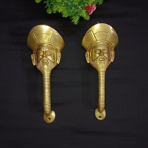 May include: A pair of gold-toned door handles with a detailed face design. The handles are made of metal and have a curved shape.