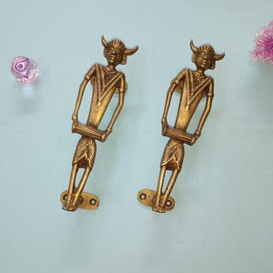 May include: Two gold-toned metal door handles shaped like figures playing a musical instrument. The figures are holding a drum-like instrument in their hands. The handles are mounted on a light blue background.