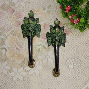 以下が含まれることがあります： 緑と金の仕上げが施された装飾的なドアノブが2つ。ノブは、王冠と翼のある顔の形をしています。ノブは白い背景に設置されています。