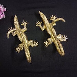 Pode incluir: Dois ganchos de parede em forma de lagarto de cor dourada. Os lagartos estão virados em direções opostas e têm escamas e garras detalhadas.