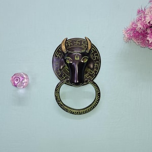 Op de afbeelding: Een messing deurklopper met een stierenkopontwerp. De stierenkop is zwart met gouden accenten en de deurklopper heeft een ronde basis met een swastikapatroon.