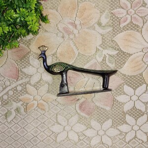 May include: A decorative peacock figurine, primarily black with green and gold accents. The peacock has a long, curved body and a detailed tail. The figurine is made of metal and sits on a small base. The background is a floral patterned fabric.