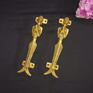 May include: A pair of gold-toned door handles shaped like women with their hands clasped in prayer. The handles have a detailed design and a fishtail base.