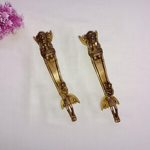 Peut inclure: Deux poignées de porte en métal doré avec des motifs complexes représentant des figures humaines. Les poignées sont longues et fines avec une forme décorative en forme d'aile en bas.