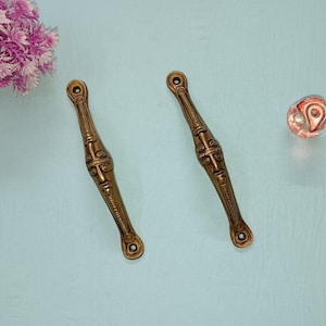 May include: Two ornate brass cabinet door pulls with a face design. The pulls are on a light blue background.