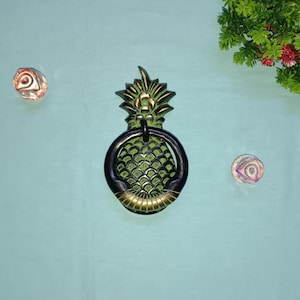 Puede incluir: Un llamador de puerta de piña verde y dorado con un anillo negro. El llamador está sobre un fondo azul claro.
