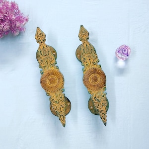 Puede incluir: Dos manijas de puerta alargadas con un acabado metálico dorado y verde oscuro con intrincados patrones florales y de hojas en relieve. Cada manija tiene un diseño central circular prominente con líneas radiantes y extremos puntiagudos.
