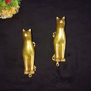 May include: Two gold-colored metal cat figurines. The cats are standing upright with their tails curled around their legs. They have a detailed design and a shiny finish.