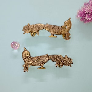 May include: Two ornate brass peacock-shaped drawer pulls. The pulls are detailed with engraved patterns and have a warm, golden hue. The peacock's tail feathers extend to the left, and the head faces right. A small, round base supports each pull. A pink flower and a glass ball are in the background.