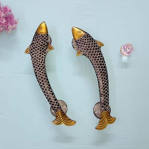 May include: A pair of decorative door handles shaped like fish. The handles are black with a scale pattern, and gold accents on the head and tail. They are curved and mounted on a light blue surface.