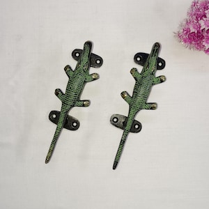 Peut inclure: Deux poignées de porte en forme d'alligator vert avec des accents en métal noir. Les poignées sont montées sur un fond blanc.