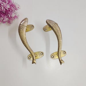 Puede incluir: Dos tiradores de puerta en forma de pez de color dorado con un acabado plateado en la cabeza y la cola. Los tiradores están montados sobre un fondo blanco.