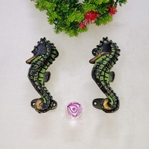 May include: Two seahorse-shaped cabinet pulls with a distressed black and green finish. The seahorses have a bronze-colored beak and base. A small, round, purple glass knob is also present.