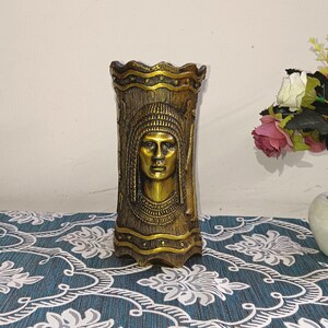 Könnte beinhalten: Eine goldfarbene Metallvase mit einem dekorativen Design, das ein Reliefgesicht zeigt. Die Vase hat eine strukturierte Oberfläche und einen gewellten Rand.