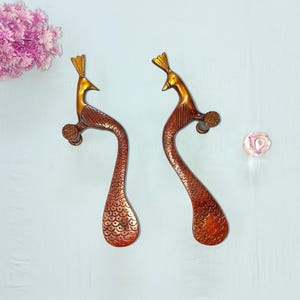 Puede incluir: Dos tiradores de puerta decorativos con forma de pavo real. Los tiradores son de metal color bronce con detalles dorados. Tienen forma curva y presentan un detallado diseño de plumas. Diseñados para ser montados en una puerta.