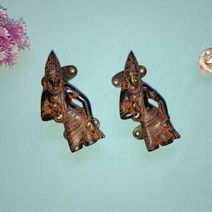 Könnte beinhalten: Zwei verzierte, dunkelbraune Türklinken aus Metall mit aufwendigen Designs, die eine Figur in einem fließenden Gewand zeigen. Die Griffe sind auf einer hellblauen Oberfläche montiert.
