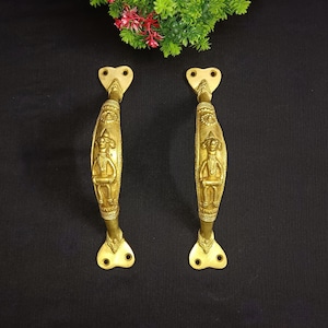 May include: A pair of gold-toned door handles with a decorative design featuring a figure playing a musical instrument. The handles have a heart-shaped top and a curved design.