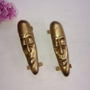 May include: Two gold-toned metal door handles shaped like stylised human faces with closed eyes. The handles have a smooth, polished finish and are mounted on a white background.