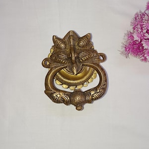 Peut inclure: Un heurtoir de porte en or avec un design détaillé d'une créature mythique à la bouche grande ouverte. Le heurtoir est en métal et possède une bague pour tirer.