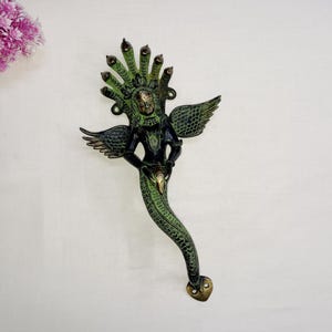 May include: A green and gold metal door handle shaped like a mythical creature with wings and a snake-like tail. The creature has a human-like face and is holding a small object in its hand.