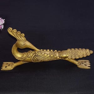 Może przedstawiać: Złoty, dekoracyjny przedmiot w kształcie pawia. Ptak ma bogaty wzór z detalami piór i długą, zakrzywioną szyję. Na ciemnym tle, z różowymi kwiatami w lewym górnym rogu.