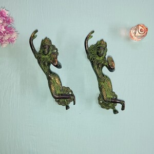 Op de afbeelding: Een paar sierlijke messing deurgrepen in de vorm van een dansende vrouw. De grepen zijn groen en goud van kleur en hebben ingewikkelde details.